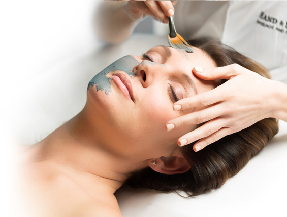 A woman's face with her eyes closed and someone behind her in a Hand & Stone smock applying a mask to her face with a brush